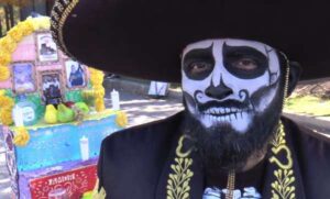 A man with his face painted as a skull poses in front of an ofrenda during the Mexican celebration of the Day of the Dead